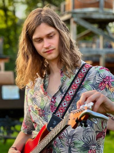 Gitarrist Anton im Porträt stimmt seine Gitarre, er trägt lange Haare und hat ein Hemd mit Blumenmustern an