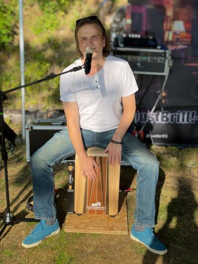 Dirk sitzt auf seinem Cajon, das hellbraun ist und mit einem holzfarbenen Streifen versehen, er trägt Jeans und ein weißes Shirt